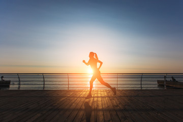 Silhouette of a young beautiful athletic girl with long blond hair in headphones, who listens to music and runs at dawn over the sea
