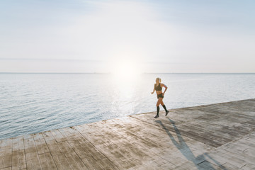 Young beautiful athletic girl with long blond hair in headphones listening to music and running at sunrise over the sea