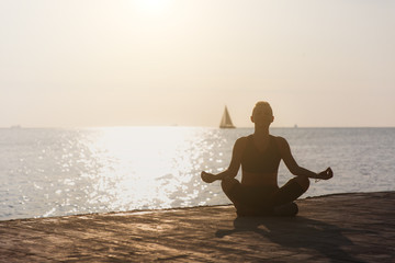Silhouette of a young athletic girl who is engaged in yoga at sunrise on the beach with a sailboat