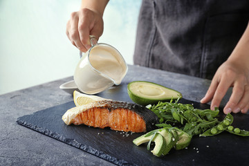 Woman pouring sauce onto slate plate with delicious salmon in kitchen