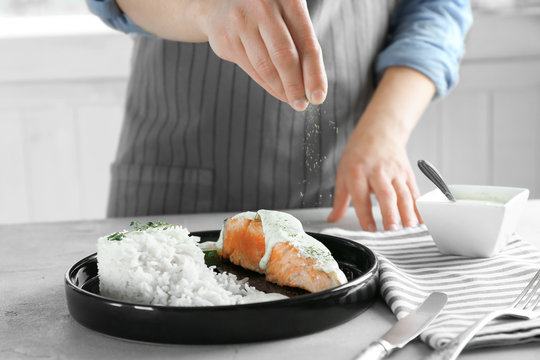 Woman adding spices to delicious salmon and rice on plate in kitchen