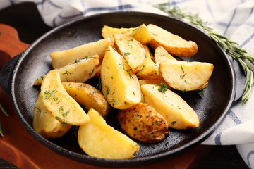 Frying pan with delicious baked potato wedges on wooden board, close up