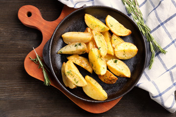 Frying pan with delicious baked potato wedges on wooden board