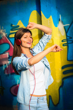 Beautiful woman listening to music on red headphones sings and dances on graffiti background 
