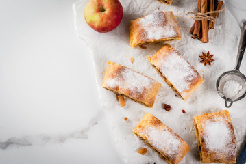 Home autumn, summer baking, puff pastries. Apple strudel with nuts, raisins, cinnamon and powdered sugar. On white marble table. Sliced, with ingredients. Copy space top view