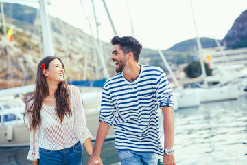 Attractive couple exploring on their summer vacation laughing as they hold hands while sightseeing a small boat harbour on the coast