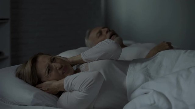 Man snoring in bed, woman beside him pressing hands against ears, troubled sleep