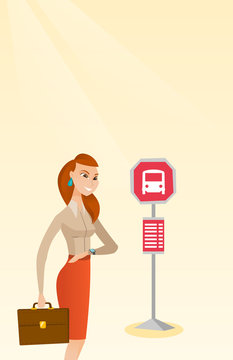 Caucasian business woman waiting for a bus at the bus stop. Young cheerful woman standing at the bus stop. Woman looking at her watch at the bus stop. Vector cartoon illustration. Vertical layout.