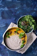 Mushroom cream soup in black ceramic bowl served with turmeric powder, forest mushrooms, greens on textile napkin over dark blue texture concrete background. Top view with copy space