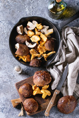 Heap of fresh forest porcini boletus and chanterelles mushrooms in iron cast pan with olive oil and salt, served won textile napkin with knife over gray texture background. Top view with space.
