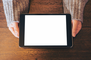 Mockup top view image of woman's hands holding and showing black tablet pc with blank white screen on vintage wooden table