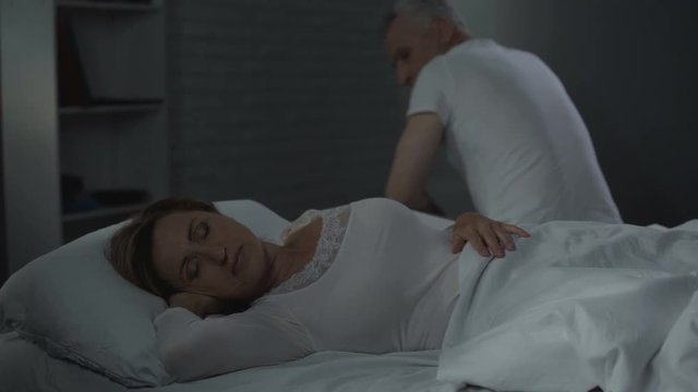 Senior woman sleeping, man sitting on far edge of bed backwards, men health