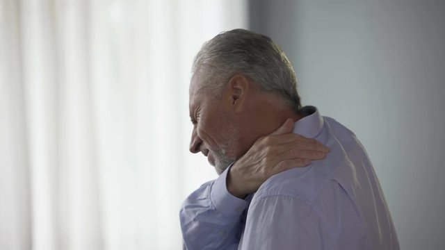 Aged man standing sideways, touching neck in acute pain, trying to move head