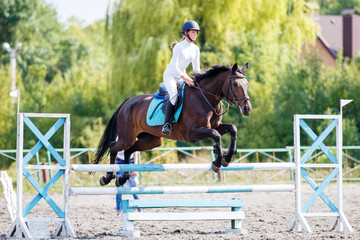 Young rider girl on bay horse jumping over barrier on equestrian sport competition. Horseback girl...