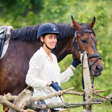 Young smiling rider woman in helmet holding bay horse by bridle