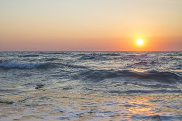 Panorama del tramonto sul mare. Il sole giallo con i suoi raggi caldi riscalda l'ambiente. Il mare è mosso e l'acqua porta a riva le alghe. Le onde increspandosi formano la schiuma.
