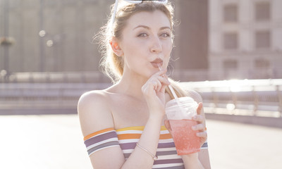 beautiful young smile blonde girl on a city street on a sunny day drinks a refreshing fruit cocktail with ice