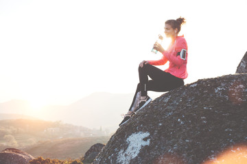 Athletic woman getting ready for a run in the mountains at sunset and drinking water. Sport tight clothes.