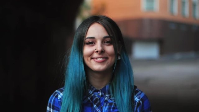 Street punk or hipster girl with blue dyed hair. Woman with piercing in nose, violet lenses, ears tunnels and unusual hairstyle stands in city. Slow motion.