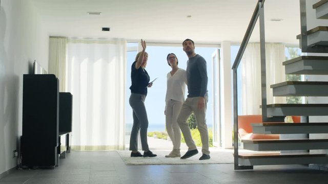 Professional Real Estate Agent Shows Stylish Modern House to a Beautiful Young Couple Who are in the Market for Purchasing/ Renting New Home. House Has Floor to Ceiling Windows and Seaside View. 