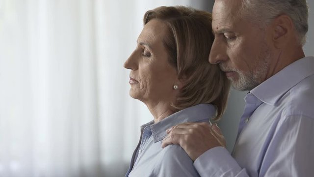 Sad elderly female standing by window, male taking her by shoulders, support