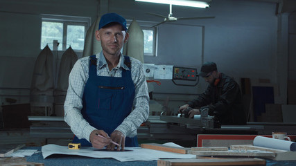 Worker using pencil while crafting furniture in factory room then turn to camera and looking.