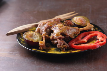 Fried meat with plums and red bell pepper on a black plate with a wooden fork on a rusty background