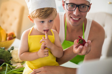 Portrait of cute little boy who sitting in cook form on a table in the kitchen near smiling father in cook form who keeping chili pepper and they looking at laptop