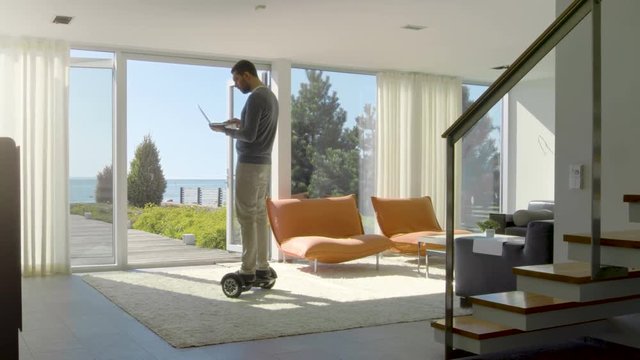 Tech Industry Businessman Working From Home Uses Laptop Computer and Rides Around the House on His Gyro Scooter. His Modern House has Seaside View. Shot on RED EPIC-W 8K Helium Cinema Camera.