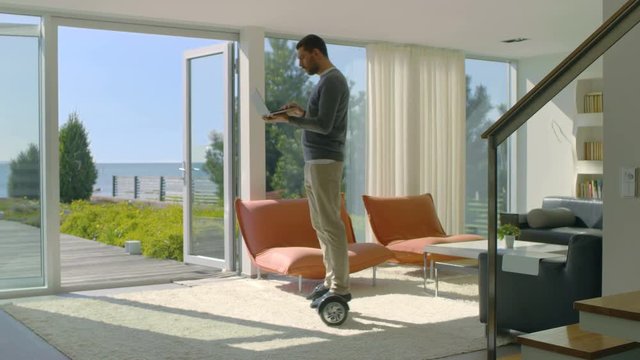 Tech Industry Businessman Working From Home Uses Laptop Computer and Rides Around the House on His Gyro Scooter. His Modern House has Seaside View. Shot on RED EPIC-W 8K Helium Cinema Camera.