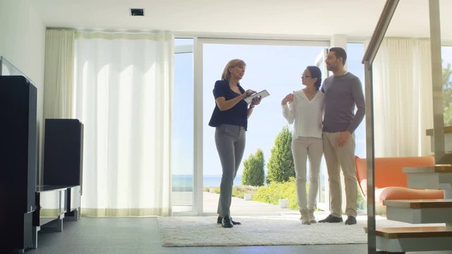 Real Estate Agent Shows Smart House Controlled with Tablet Computer to a Beautiful Young Couple. House Has Floor to Ceiling Windows and Seaside View. Shot on RED EPIC-W 8K Helium Cinema Camera.