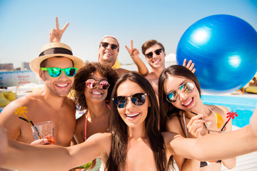 Selfie time! Six cheerful friends in fashionable different swim wear and sun protecting eyewear, caps, with drinks are posing for a selfie photo, that brunette lady is taking, beaming smiles, sunshine