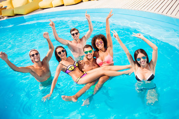 Chilling, dancing international youth, enjoying on resort beach pool sunny disco in the water, in diverse trendy fashionable bikinies, spectacles, celebrating holiday, guy holds two ladies on hands