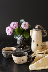 Two cups of black coffee, black chocolate, a yellow teapot and a vase with pink roses on a dark background.