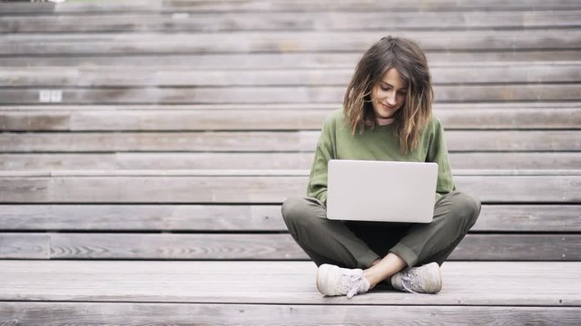 Crosslegged beautiful young woman wearing a green sweatshirt sitting on the stairs outside and typing at her laptop. Locked down real time establishing shot