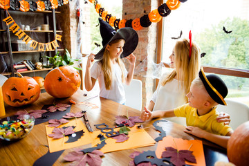 Getting ready for the halloween costume party, blond mother with two small cheerful kids, girl holds the witch hat, boy gestures like sign, bright decorated room, spooky design, bats, spider nets