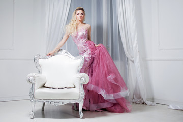Young beautiful woman in a luxurious pink dress, in interior near the white chairs