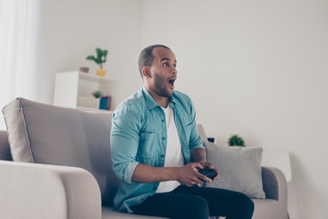 Side profile shot of shocked african young man holding joystick and playing videogames. He is very excited, sitting on couch at home © deagreez