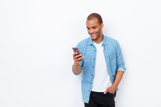 Portrait of young smiling african male student, typing sms on his phone, wearing casual jeans outfit, hand in the pocket, on pure white background