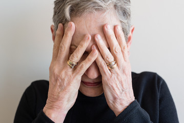 Close up of older woman in black top covering face with both hands against neutral background...