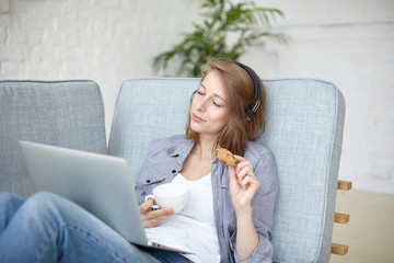 Technology and relaxation concept. Positive joyful teenage girl relaxing at home, sitting on couch with laptop, listening to favorite rock music with eyes closed, eating cookie and drinking coffee