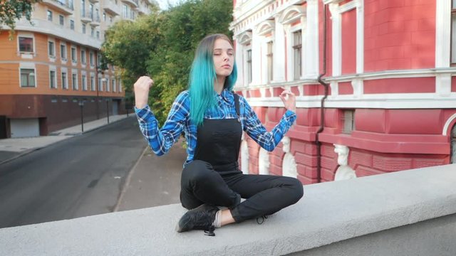 Calm street punk or hipster girl with blue dyed hair. Woman with piercing in nose, unusual hairstyle meditating on European empty street. Yoga concept. Slow motion.