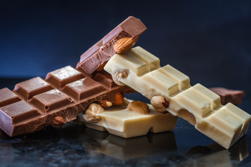Tiles of dark and white chocolate with almonds and hazelnuts on an old scratched glossy desk on a dark background