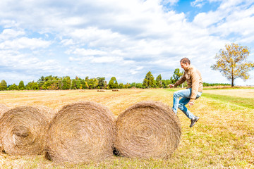 Naklejka na ściany i meble Young man jumping in air over hay roll bales in a farm rural countryside field