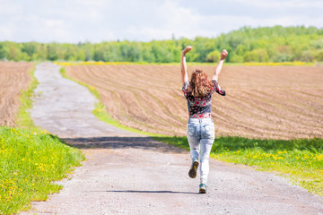 Back of young woman running, jumping in air and smiling on countryside dirt road by brown plowed...