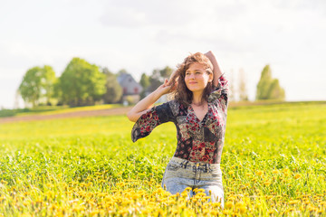 Naklejka na ściany i meble Young happy smiling woman sitting in yellow dandelion flower farm field during sunset by countryside rural house in Ile D'Orleans, Quebec, Canada raising holding, stretching hands