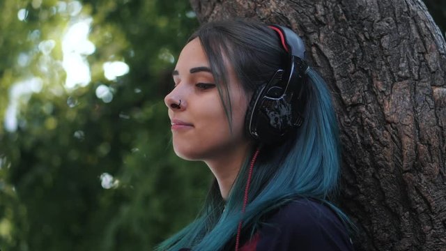 Street punk or hipster girl listening music with black headphones.Teen girl with blue colorful dyed hair,piercing in nose, sitting on big tree in park .Slow motion.