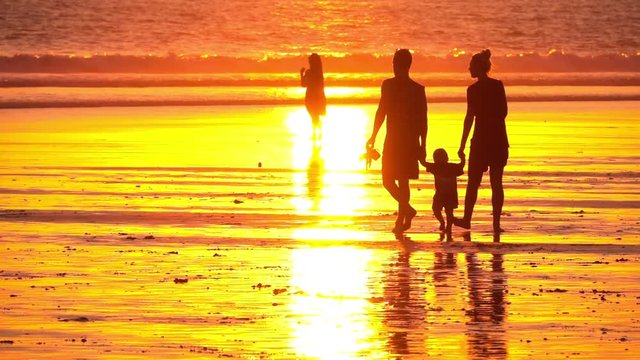 SLOW MOTION CLOSE UP SILHOUETTE: Young family with toddler walking down the smooth sandy Kuta Beach in Bali at magical golden light sunset. Mother, father and child strolling along seashore at sunrise