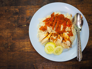 Top view Thai local traditional boiled and fried chicken rice, topped with black sweet sticky soy sauce and chili, cucumber, stainless steel fork and spoon, on dark wooden table background