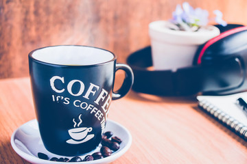 a cup of hot coffee on the brown wood table with a headphone behind for background.a black cup with coffee  word on the brown wood floor.